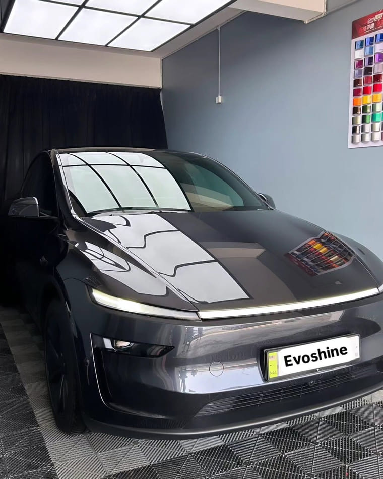 A grey Tesla Model Y seen from the front three-quarter in the bay, the ceiling panel drawn along its bonnet.